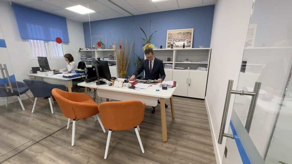 Gestor revisando documentos en su mesa de la gestoría, con una compañera al fondo ante su ordenador