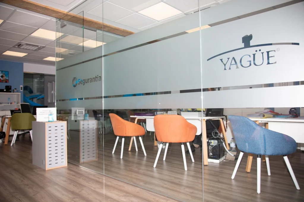 Mampara de cristal con el logotipo de Yagüe y sillas naranjas y azules para clientes en una de las gestorías de Zaragoza